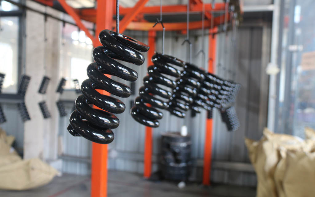 Black-coated coil springs hanging from a hook on a coating line.