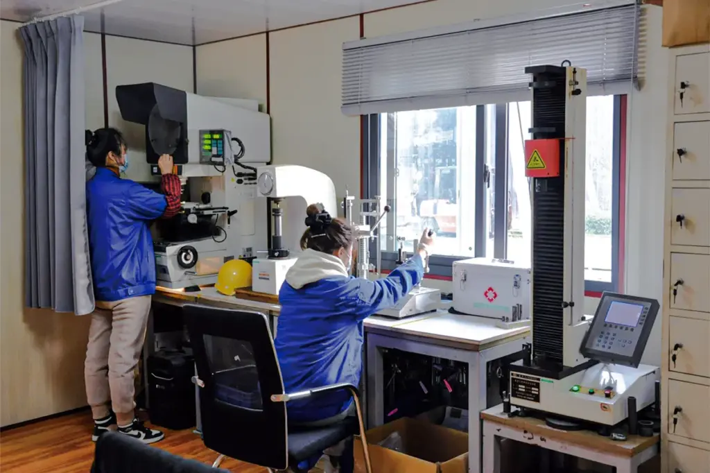 Two inspectors in the NorenSpring inspection room, one at a profile projector and one at a hardness tester, with a column-type spring load tester on the right