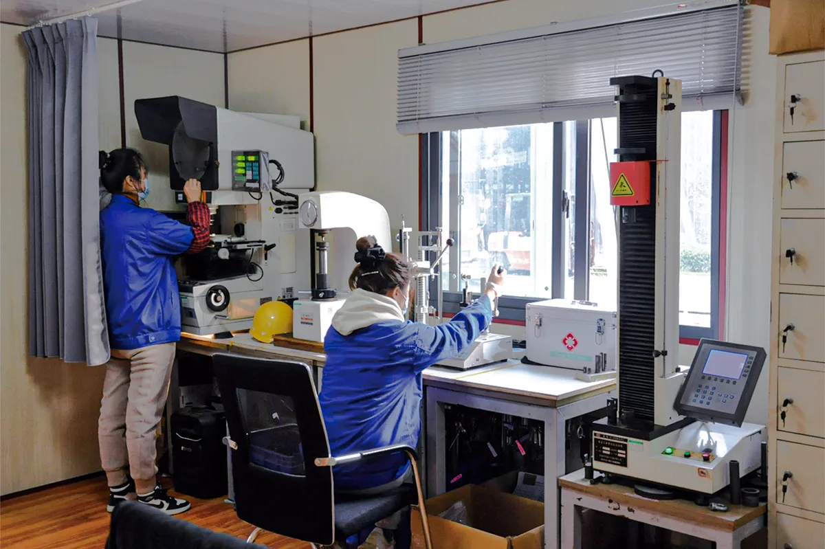 Two quality technicians in an inspection room, one at an optical measuring projector and one at a spring load testing machine