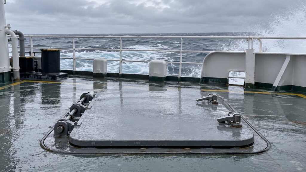 Illustration: a steel deck hatch with hinges and latches on a wet vessel deck, with rough sea behind.