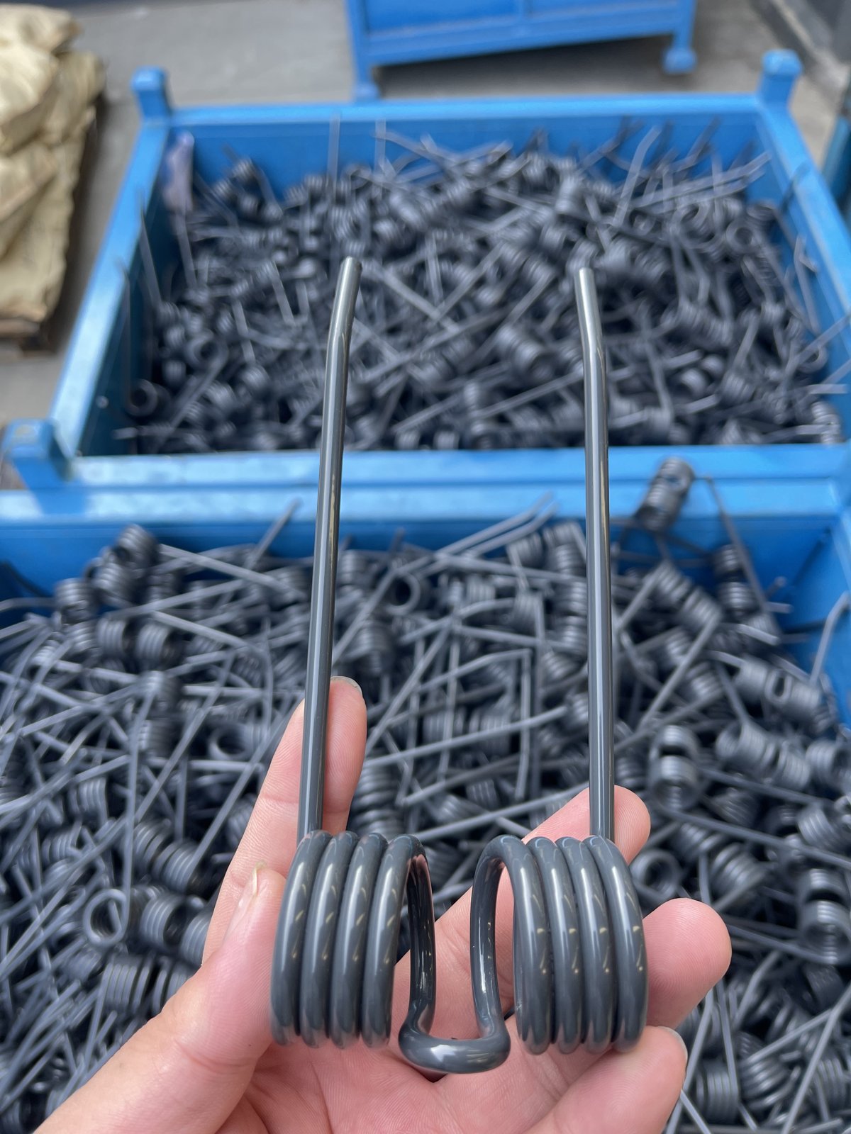 A worker's hand holding a black double torsion baler tine above steel bins filled with finished tines