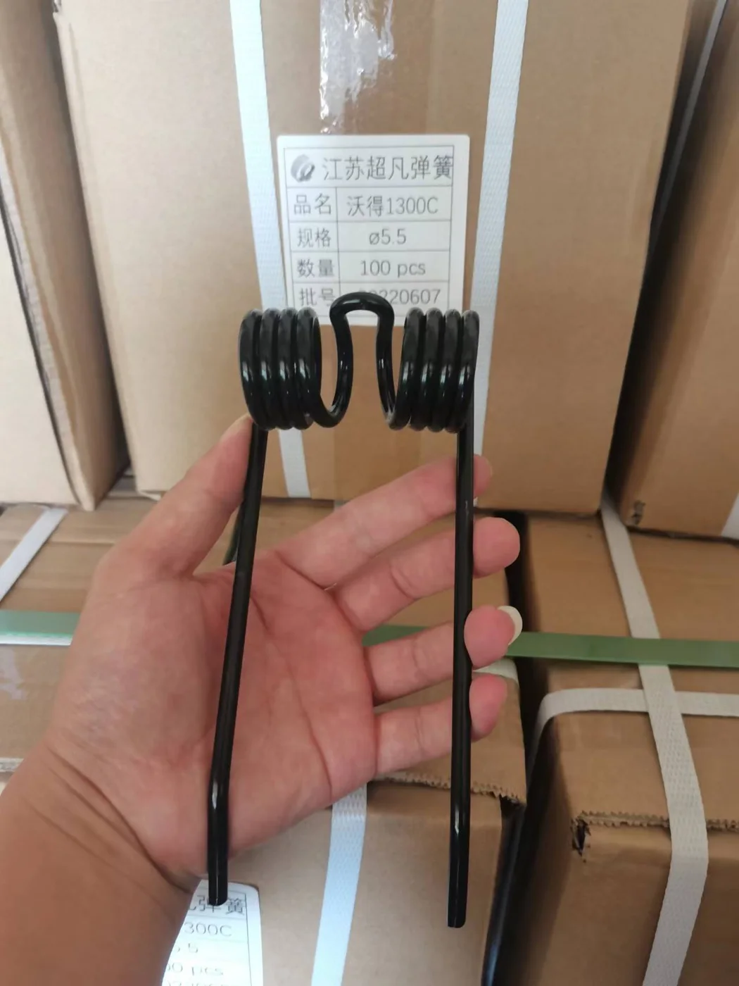 A black double torsion tine held in front of a carton label listing product name, wire size 5.5 mm, quantity 100 pcs and batch number