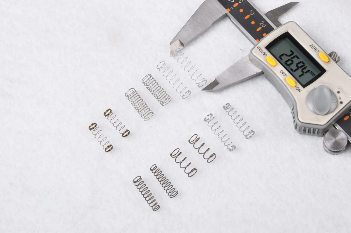 About a dozen small compression springs in stainless and plain steel laid on a white cloth beside an open digital caliper